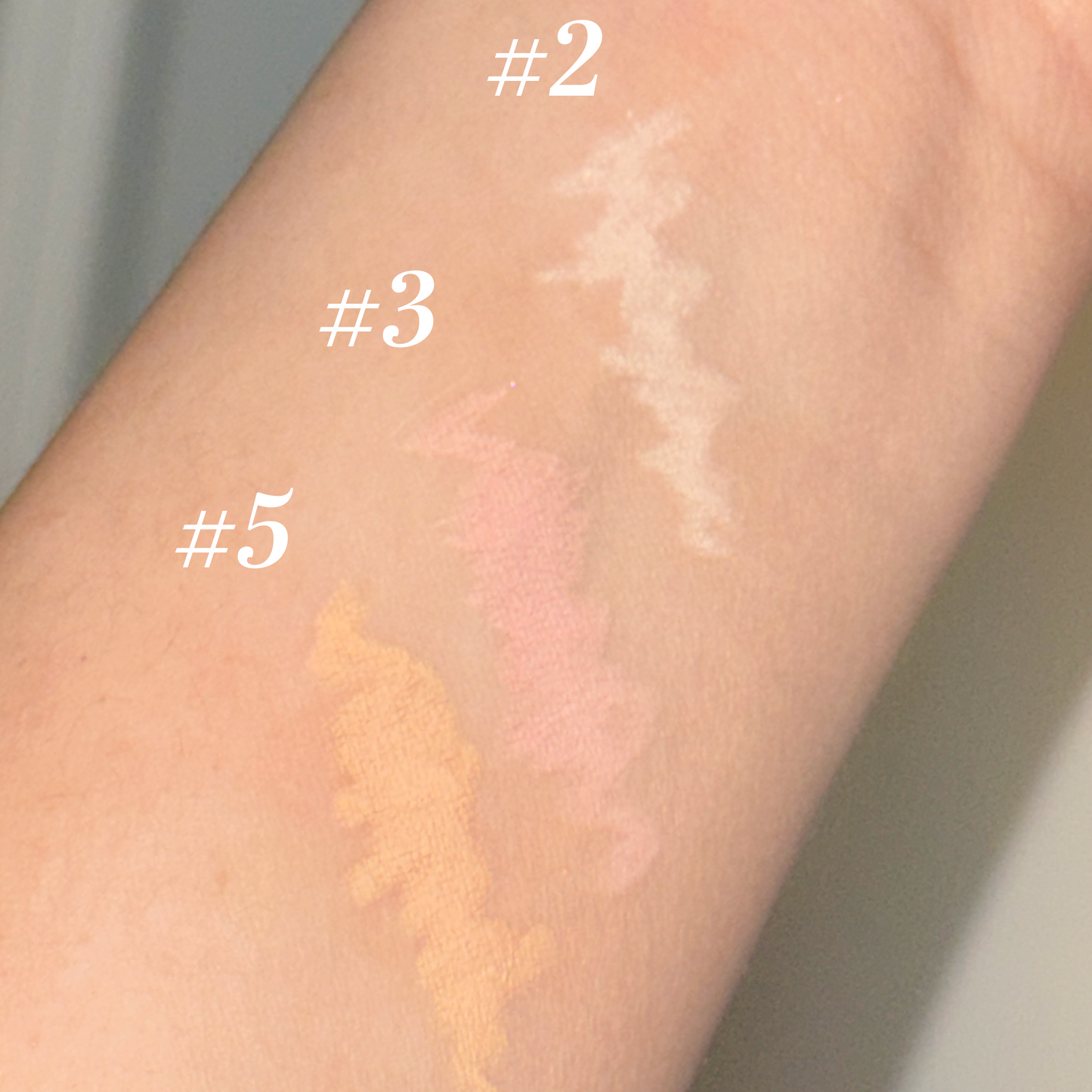 Close-up of a forearm showing three makeup swatches labeled #2, #3, and #5 in light beige, soft pink, and peachy beige tones.
