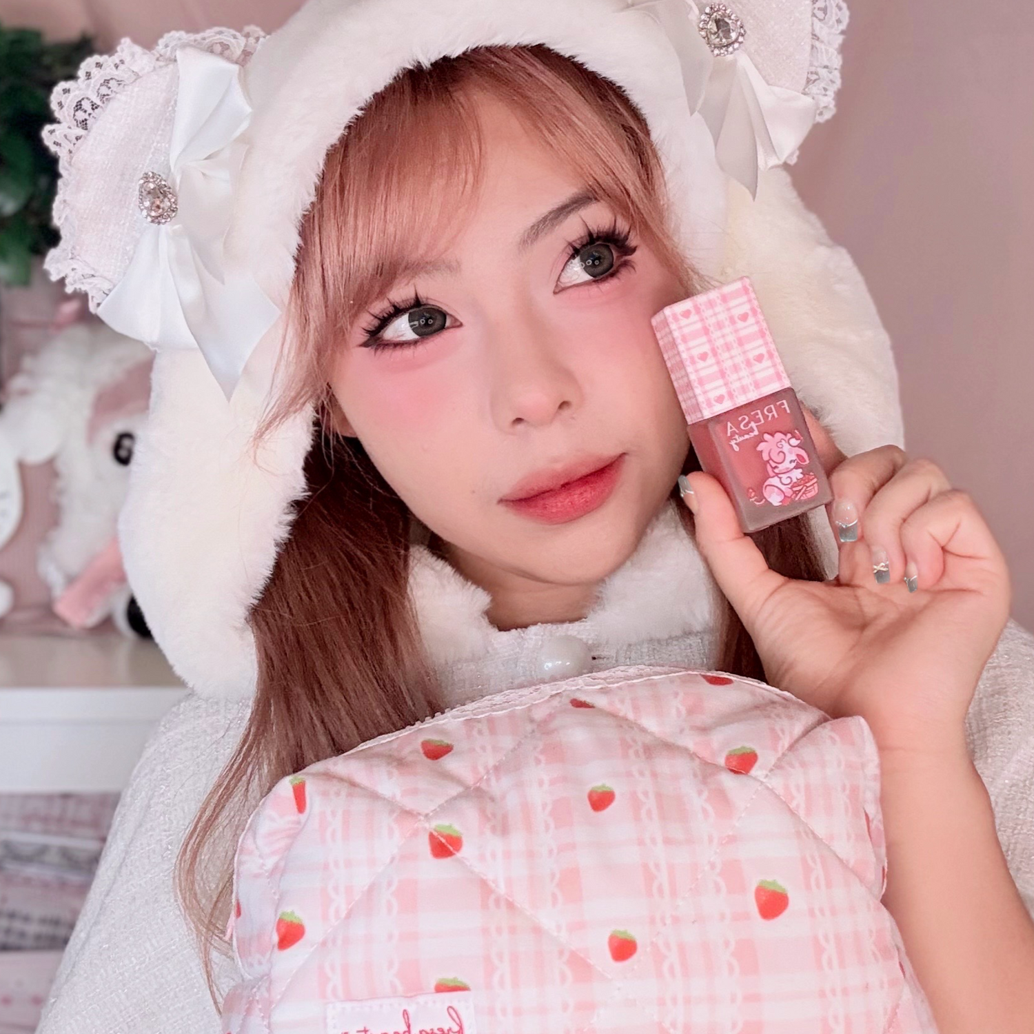 Young woman in a fluffy white hood with bows holds up a pink Fresa Beauty makeup product, featuring a cute design, against a pink plaid pillow with strawberries.