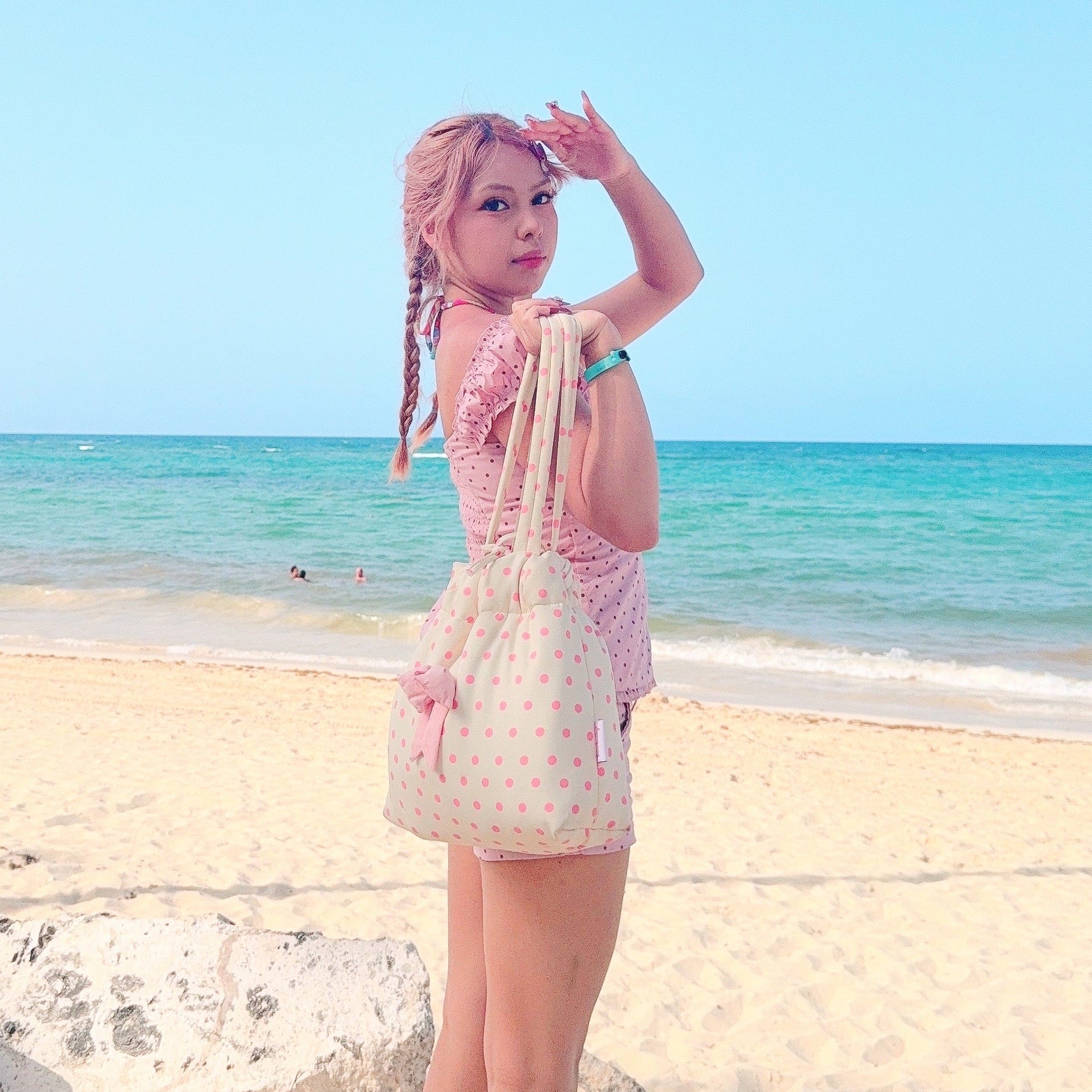 A woman stands on a sandy beach wearing a light pink romper and carrying a yellow polka dot shoulder bag with a pink bow in the middle, with the blue ocean and sky in the background.