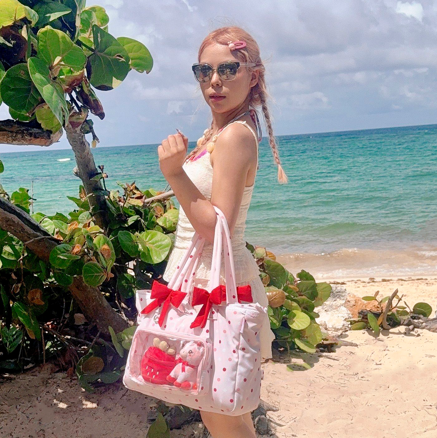 A woman on a sandy beach wearing a white dress and sunglasses, carrying a pink polka-dot tote bag with red bows and a clear front pocket containing a red plush toy. The background features turquoise ocean water, beach foliage, and a cloudy sky.