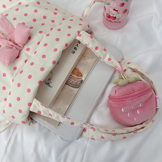 A pink and cream yellow polka dot backpack styled on white bedding with a Hello Kitty tumbler and a strawberry-shaped pink pouch inside.